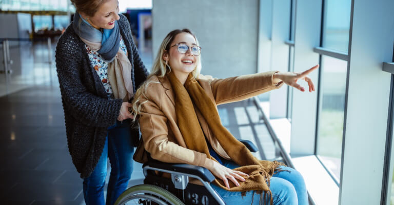 Junge behinderte Frau im Rollstuhl und ihr Begleiter lächeln in einem Flughafen und schauen hinaus.